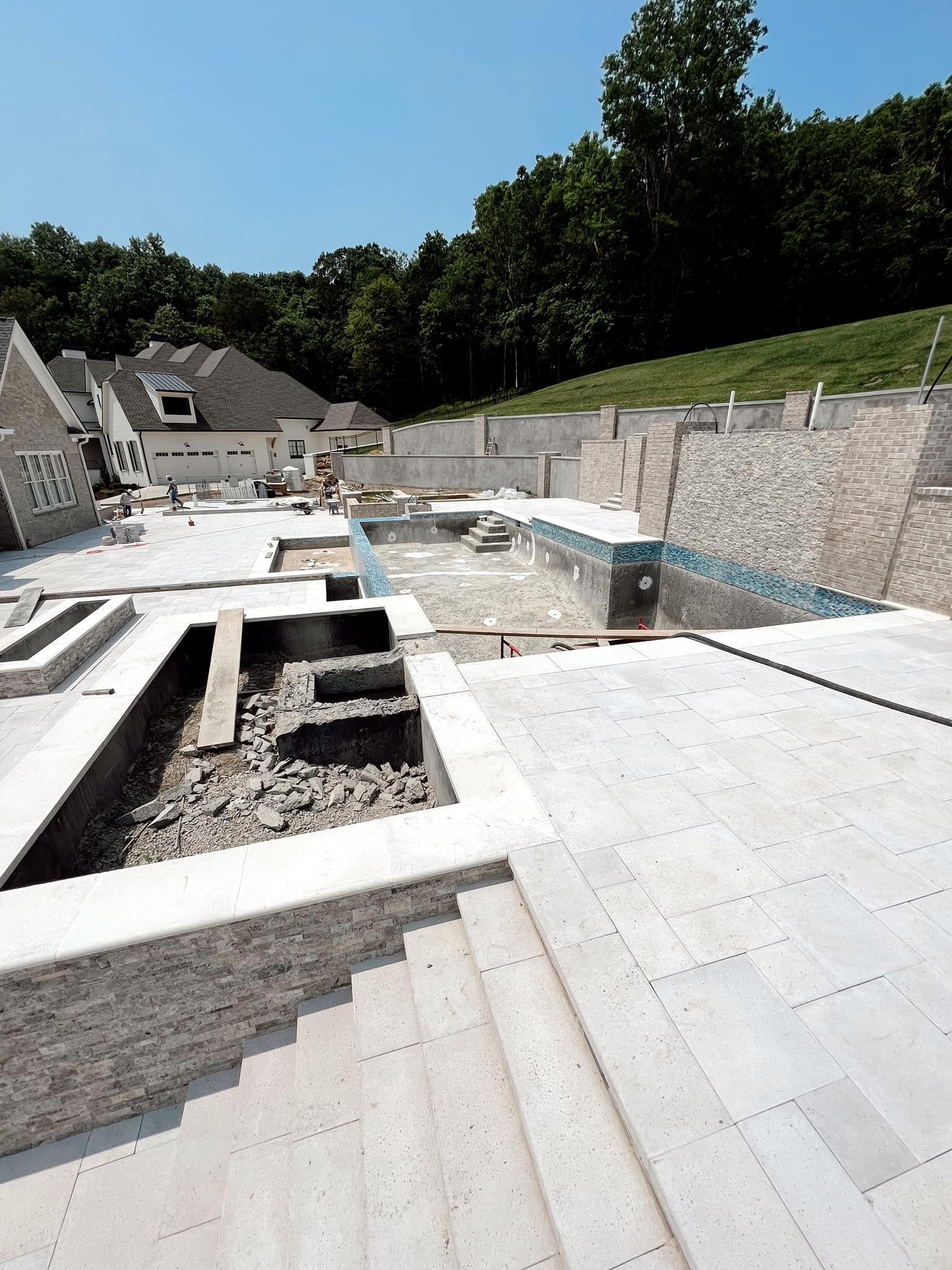 Stone pool under construction with steps, white decking, retaining walls, and a hillside backdrop