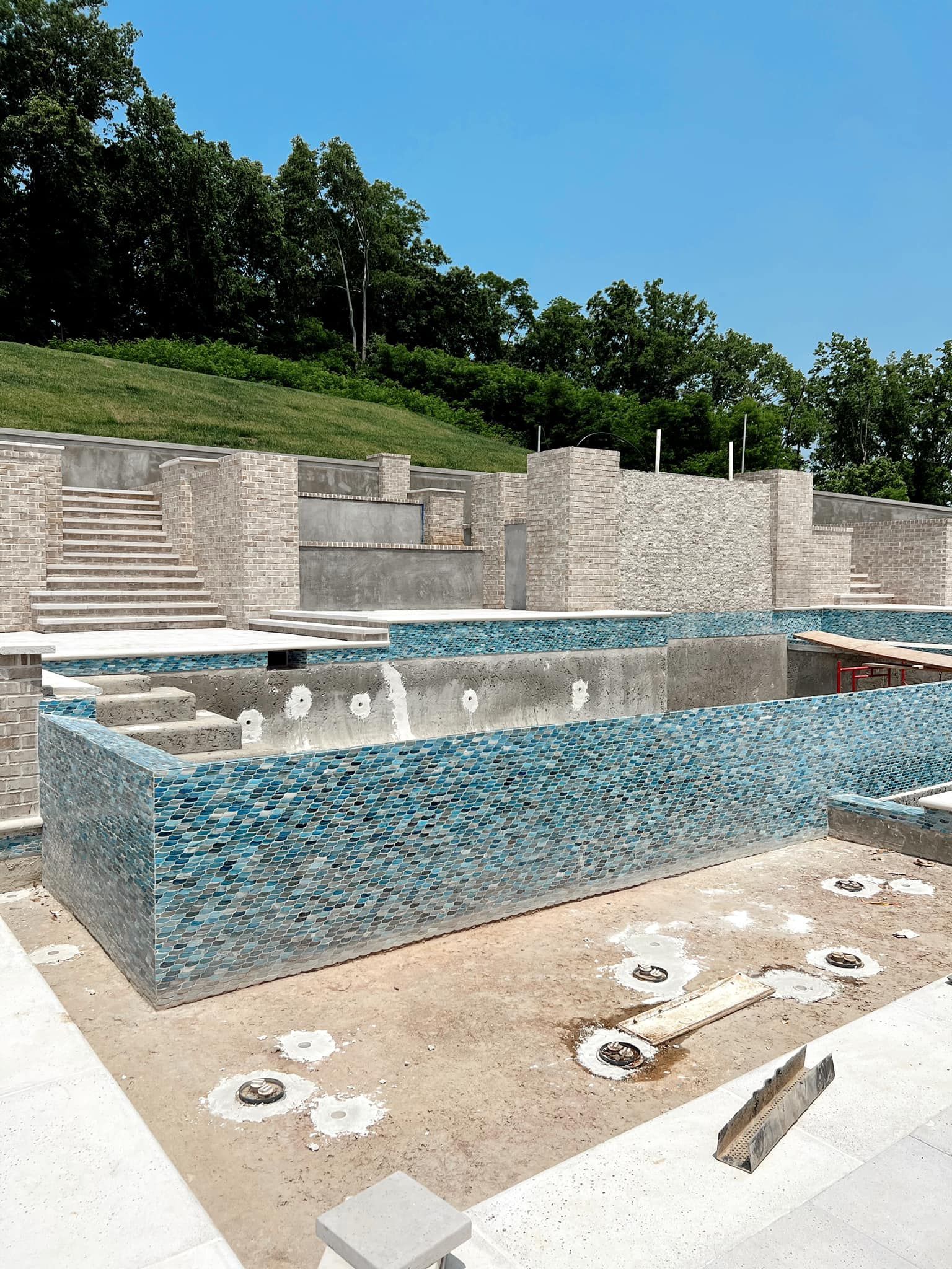 Outdoor mosaic-tiled fountain with stepped stone walls, blue water, and scattered debris in a park-like setting