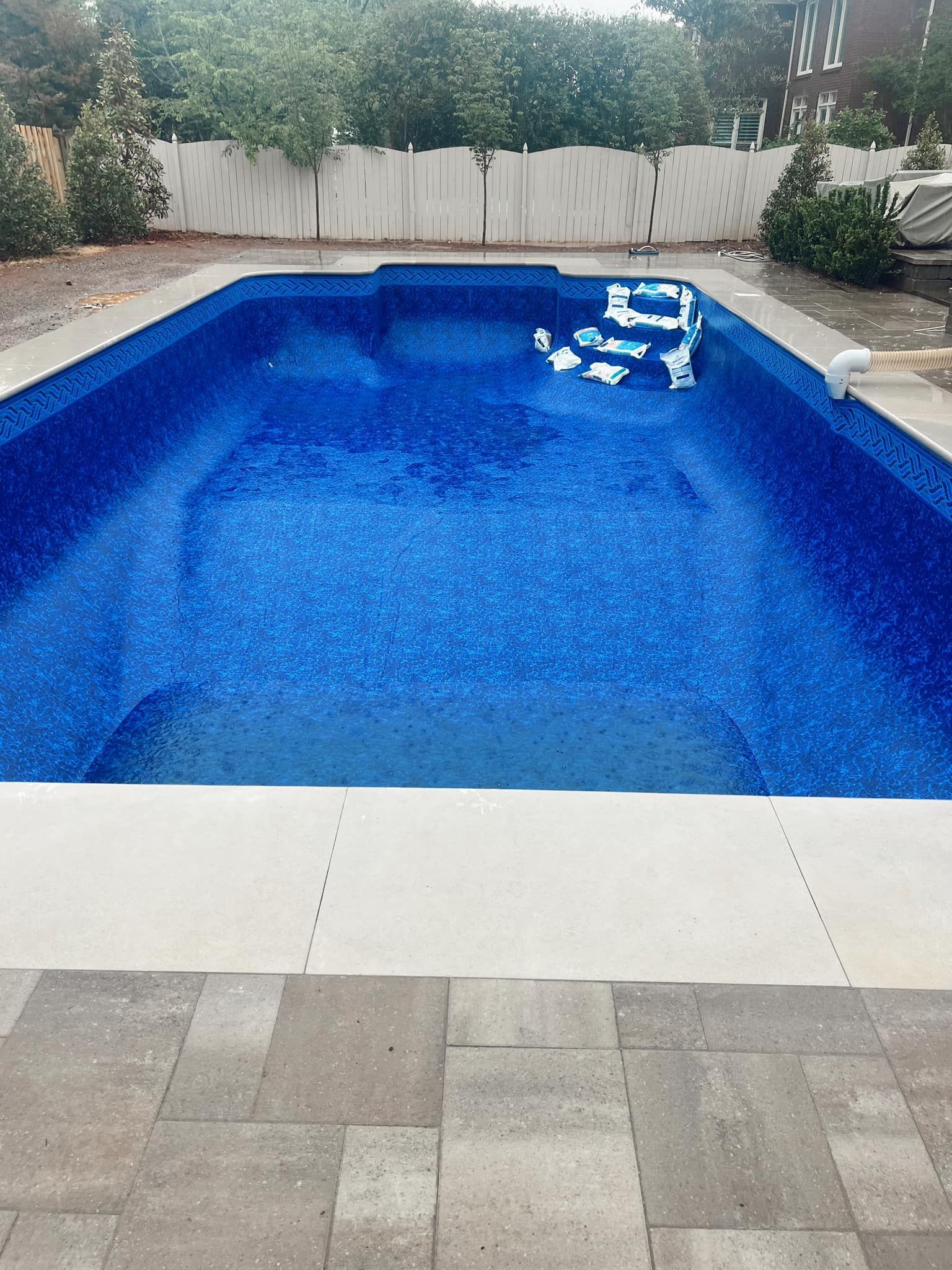 Backyard swimming pool with bright blue water and white pool steps, bordered by pavers and a wooden fence.