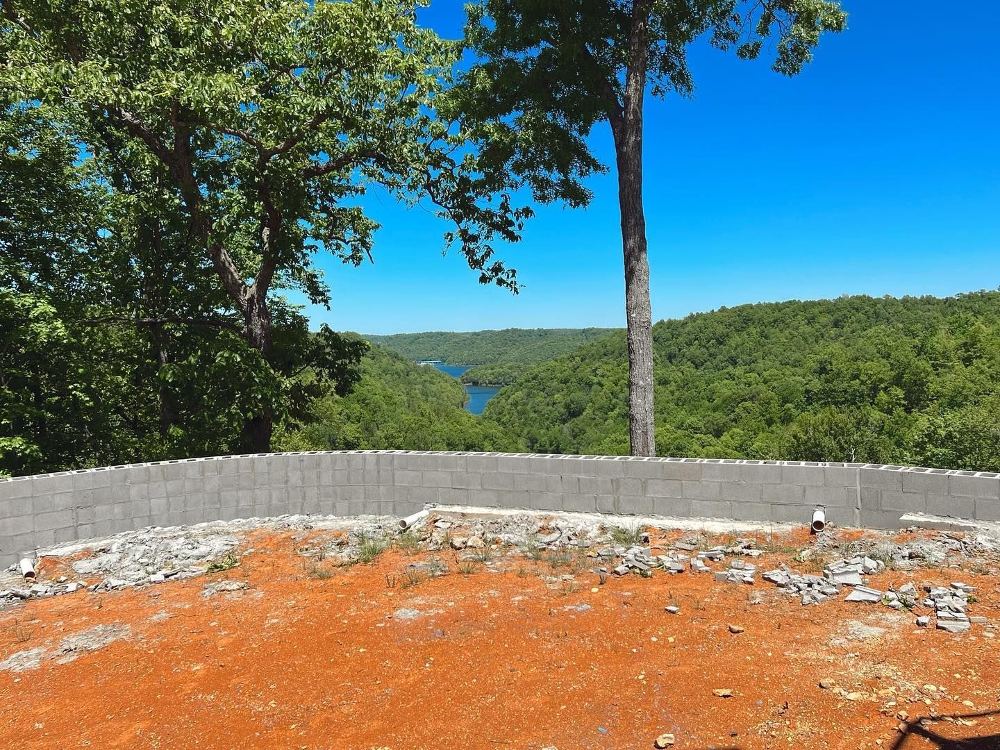 Stone overlook with orange dirt foreground, low wall, and a forested valley under a bright blue sky