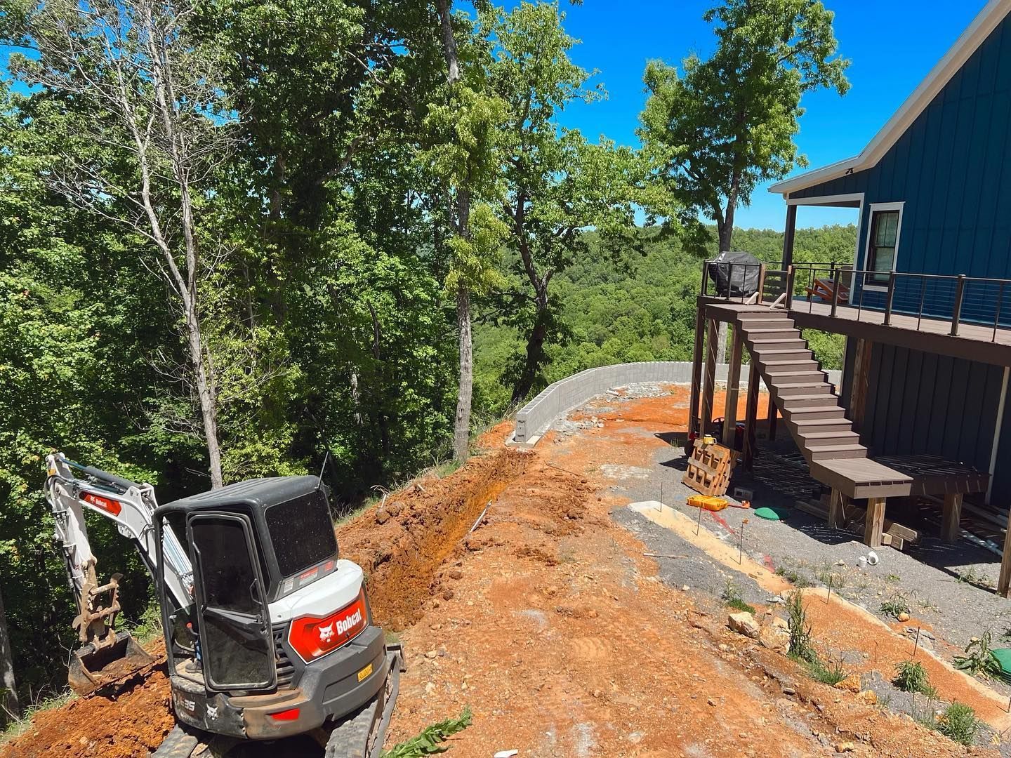 Mini excavator grading orange dirt beside a blue house on a wooded hillside.