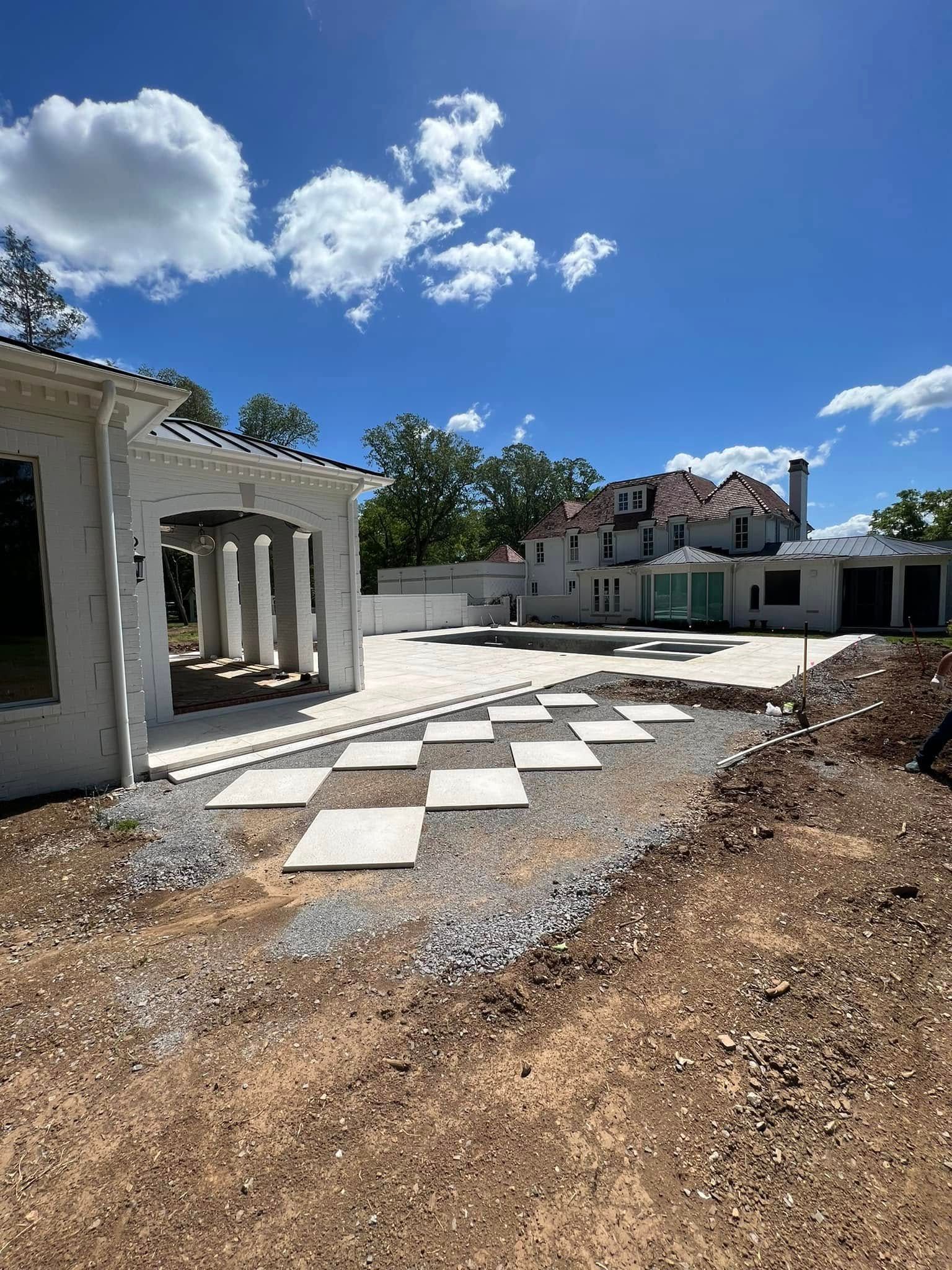 Backyard under construction with stone pavers, gravel, and a white stucco structure beneath a blue sky.