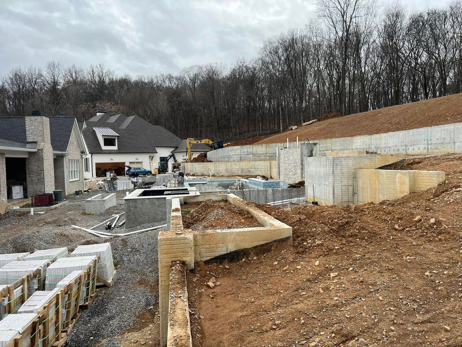 Construction site with foundation walls, gravel paths, and a partially built house on a wooded hillside