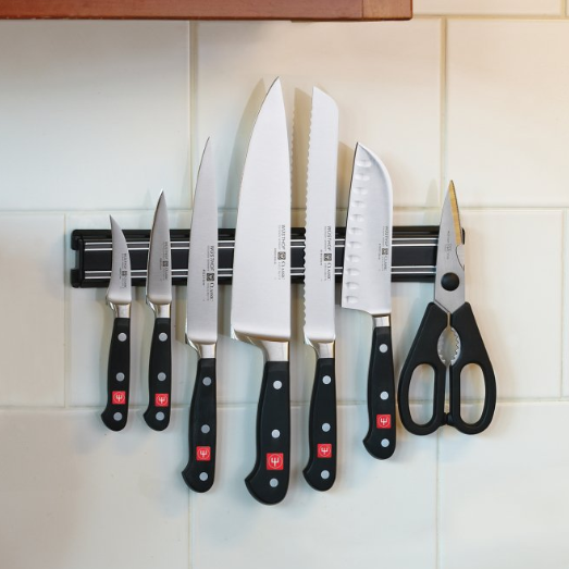 Kitchen knives and scissors attached to a magnetic strip on a white-tiled wall.