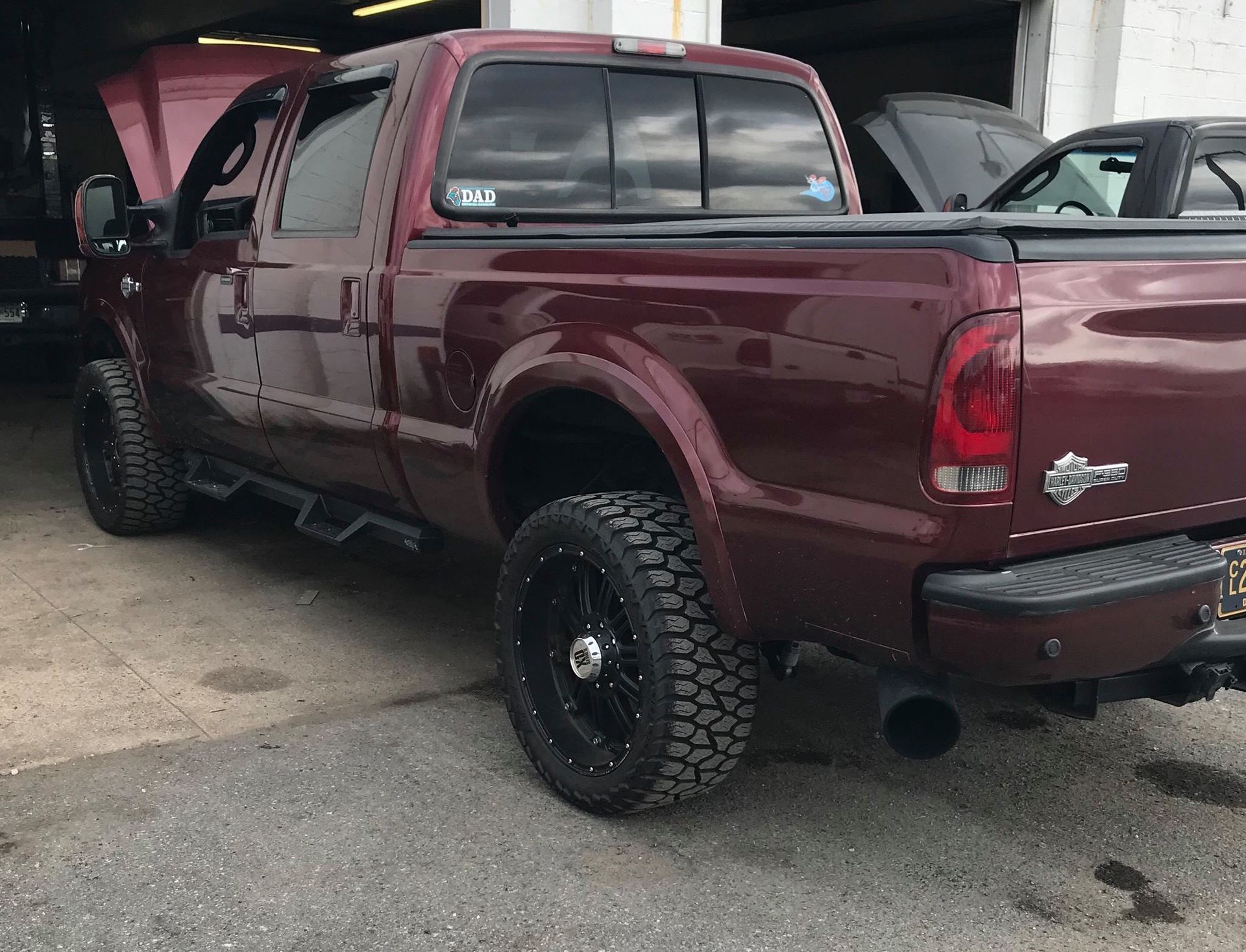 Dark red Ford pickup truck with black wheels and a side step parked in a garage.
