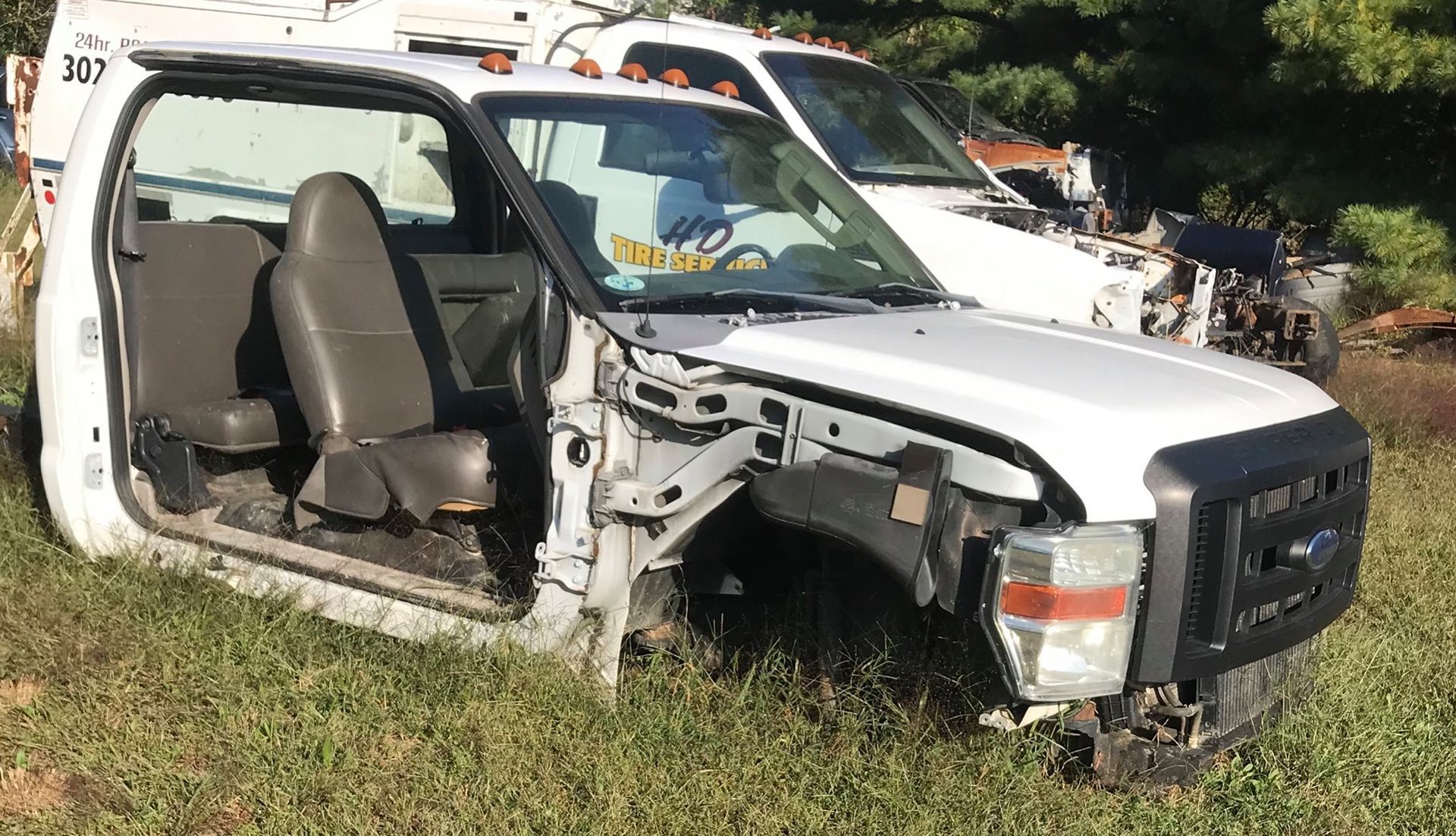 White work truck stripped of parts, parked in a grassy area, another truck in the background.