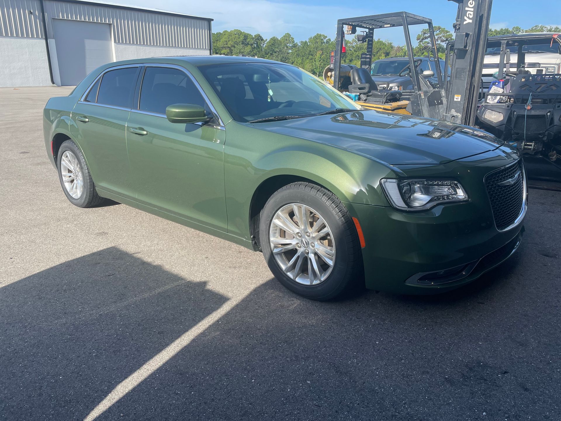 A green chrysler 300 is parked in a parking lot next to a forklift.
