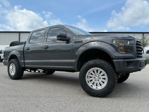 Gray lifted Ford F 150 truck with blacked out headlights and silver wheels parked on pavement under a cloudy sky.