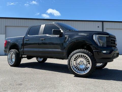 Black Ford F 150 truck with large chrome wheels, lifted suspension, parked on pavement, bright sunny day.