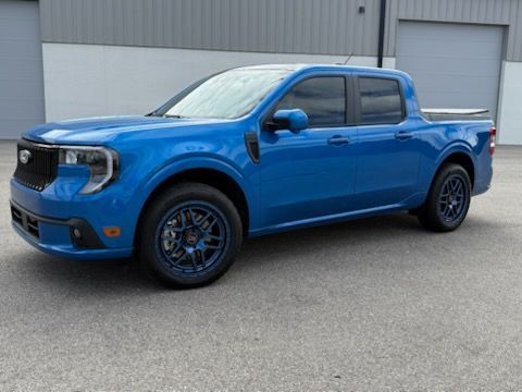 Blue Ford Maverick truck with dark blue wheels parked in front of a gray building.