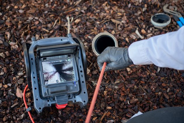 Gloved hand inspecting a pipe with a camera drain scope on a bed of mulch and debris