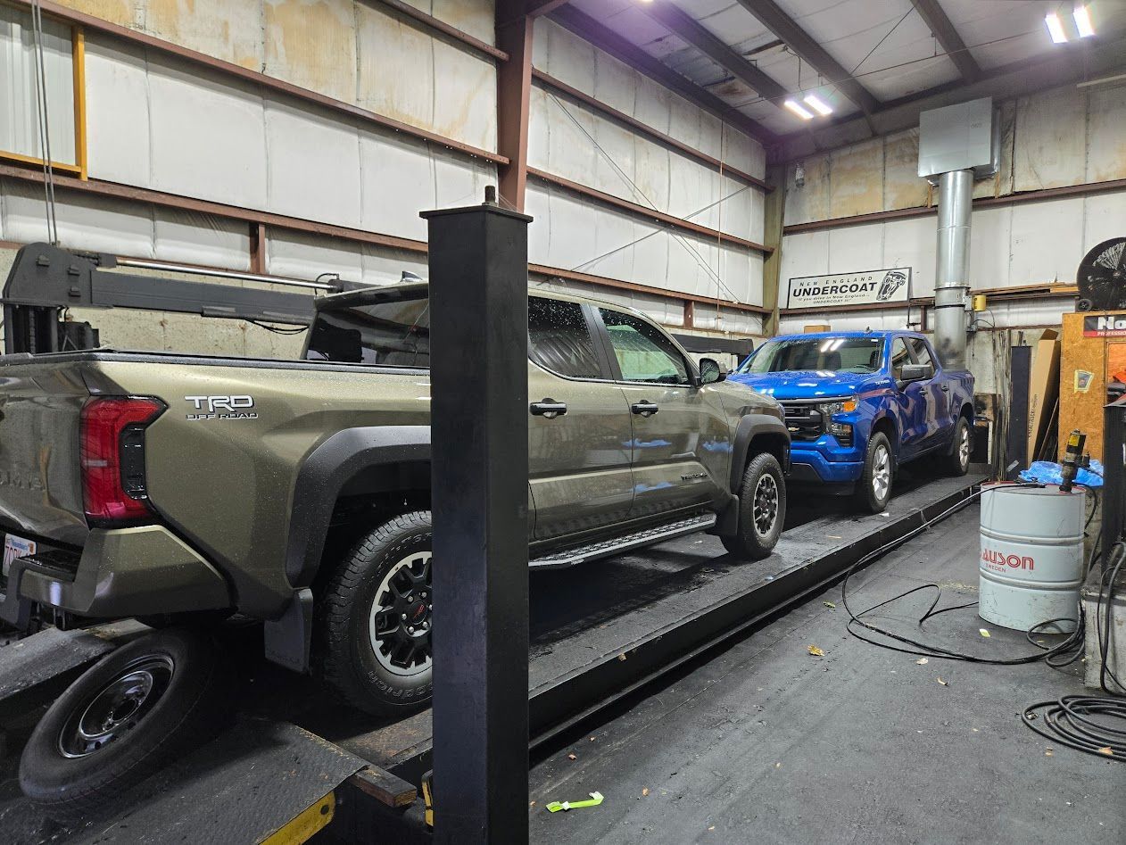 Two trucks on a lift in a repair shop. A tan one is in the foreground and a blue one is in the background.