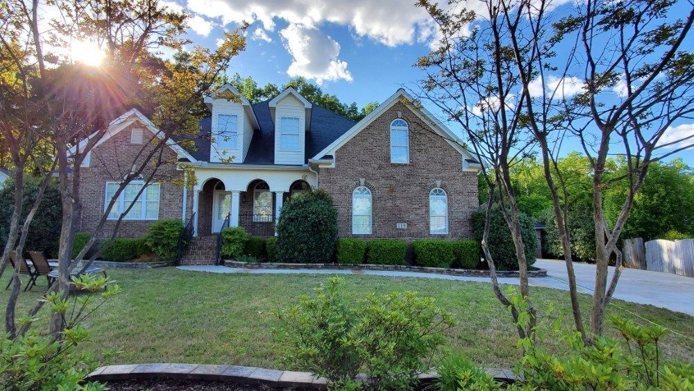 A large brick house with a black roof is surrounded by trees and bushes.