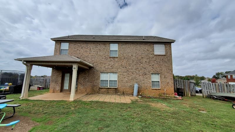 A large brick house with a porch and a lot of windows is sitting on top of a lush green field.