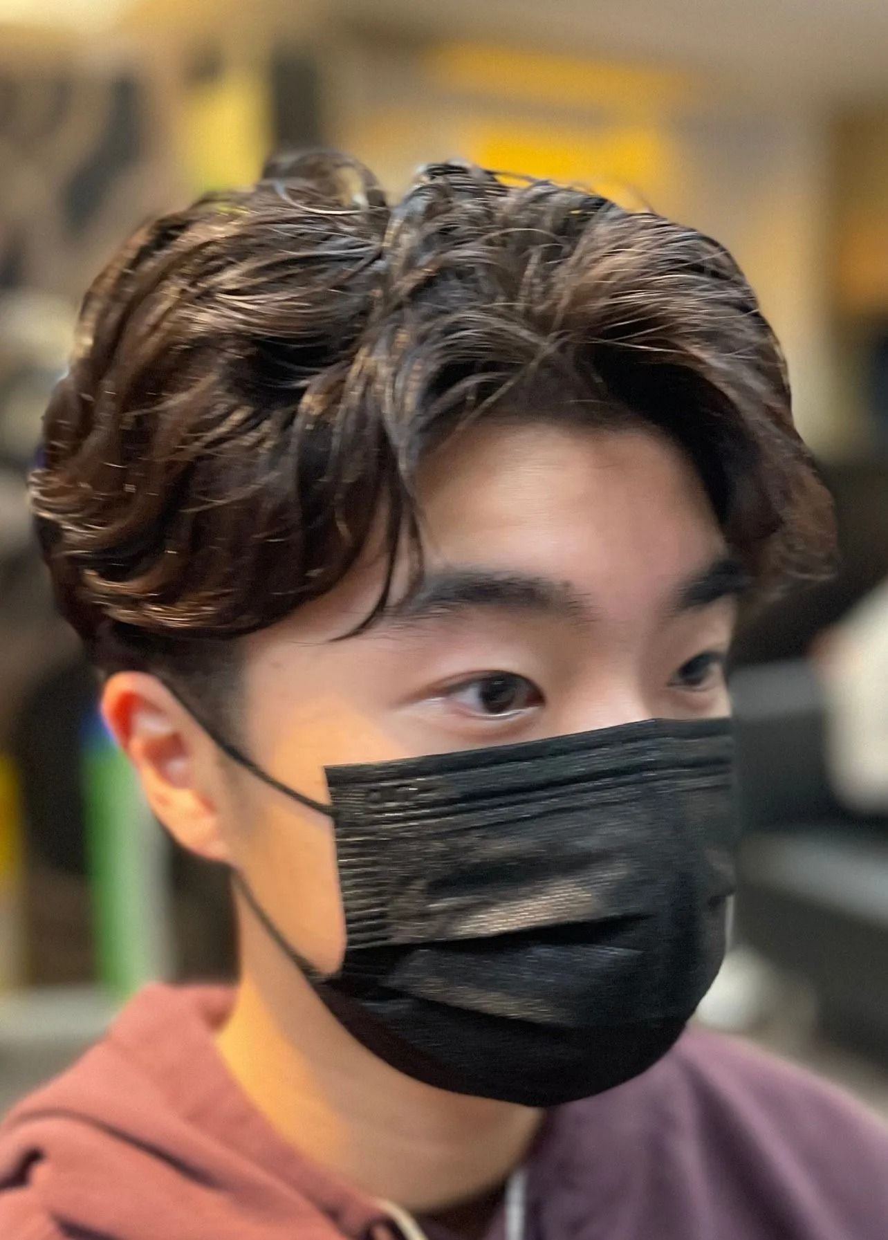 Person with dark hair and a black face mask, styled with side-swept bangs. Indoors, blurred background.