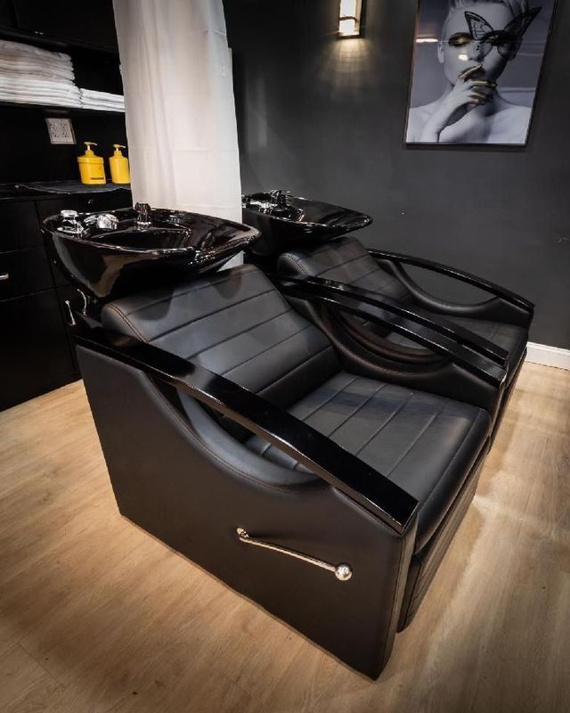 Two black salon shampoo chairs against a dark wall with a portrait and light fixtures.