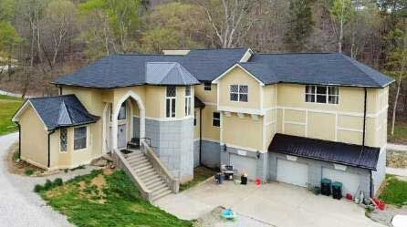 Large beige two-story house with black roof, front staircase, and attached garage on a wooded lot