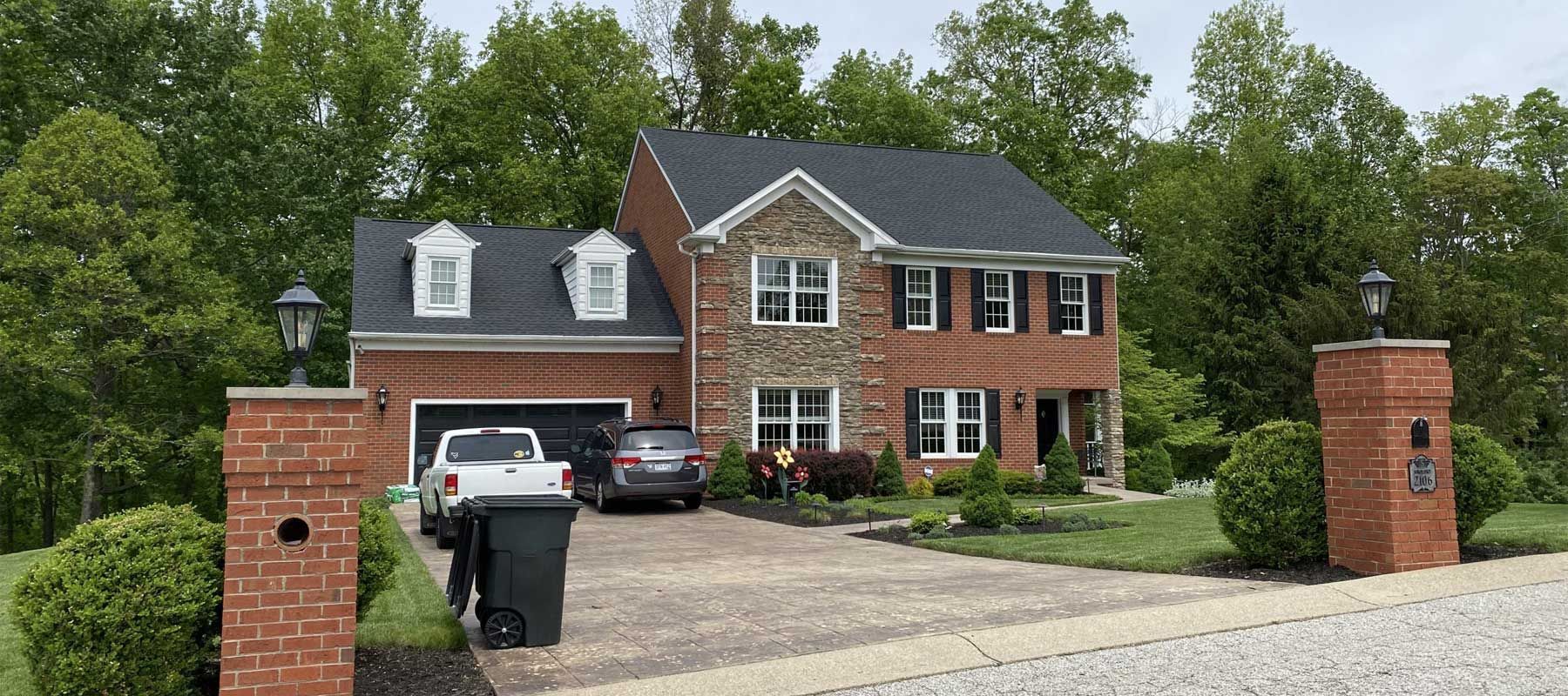 Brick suburban house with driveway, parked cars, and a trash bin by the gate