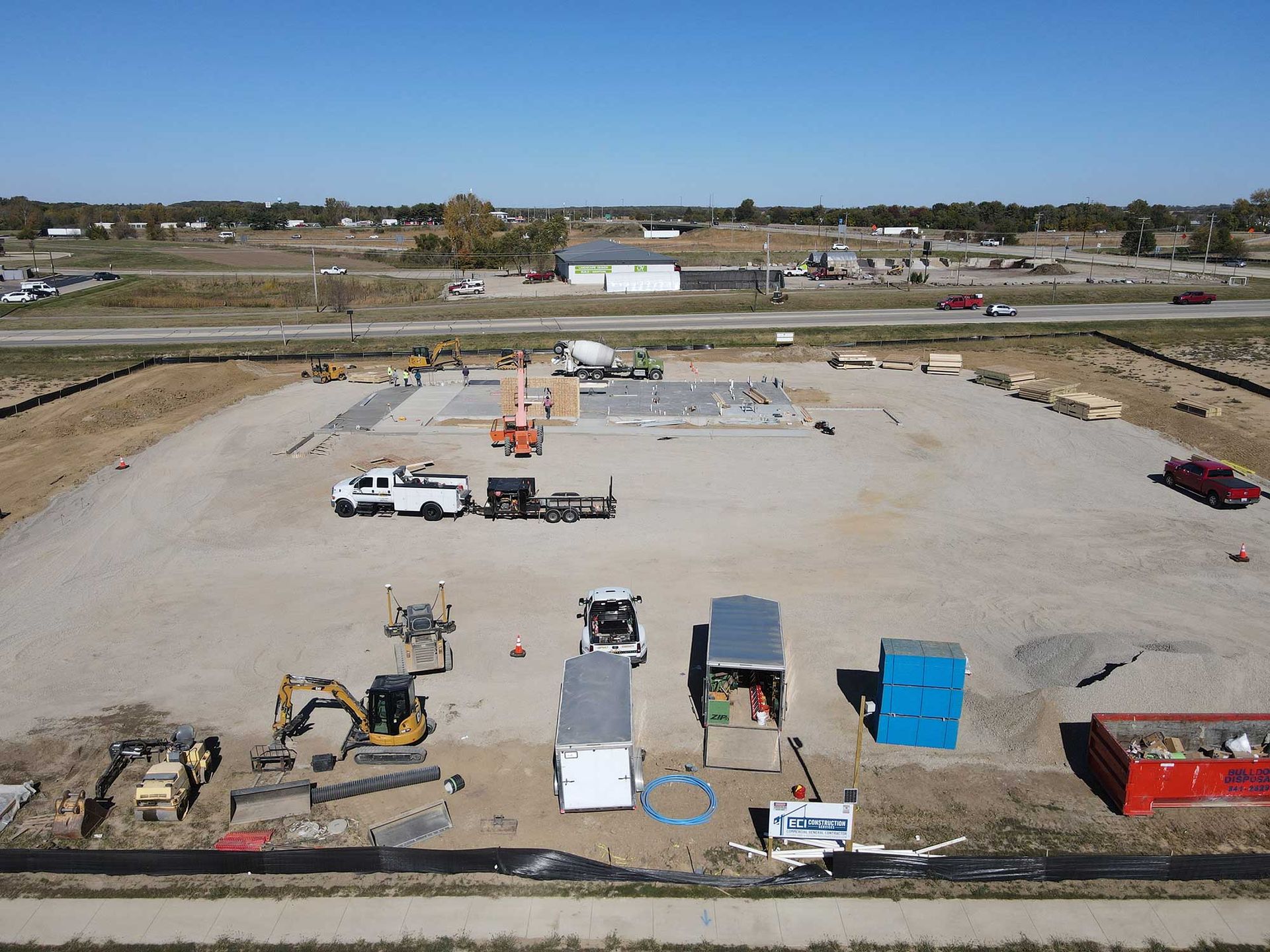 Construction site with foundation, equipment, and trailers on a gravel lot under a blue sky.