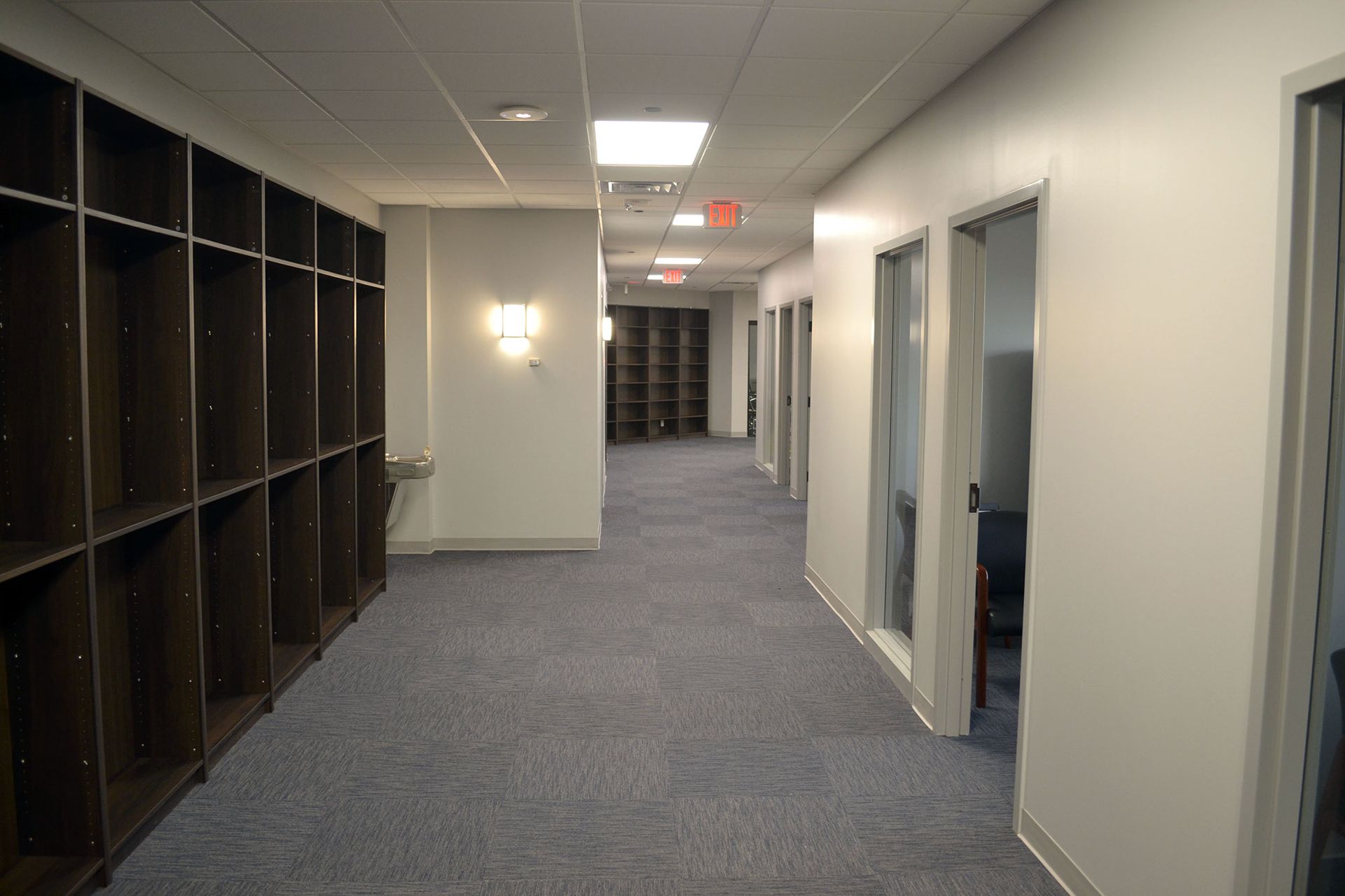 Long hallway with gray carpet, white walls, built-in shelving, and glass-walled offices.