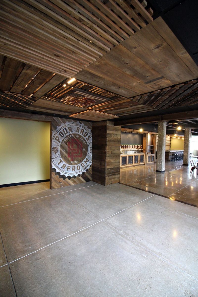 Interior of a taproom with concrete floors, wooden ceiling, logo on wall, and bar in the background.