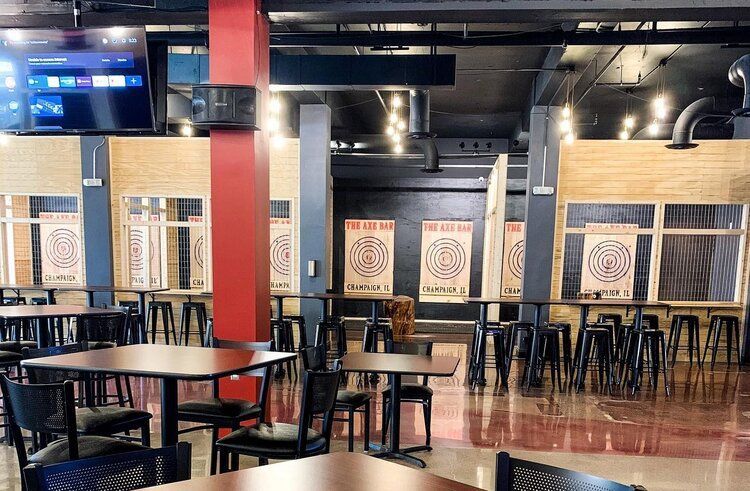 Indoor axe-throwing venue with targets, tables, chairs, and a TV. Black and red decor.