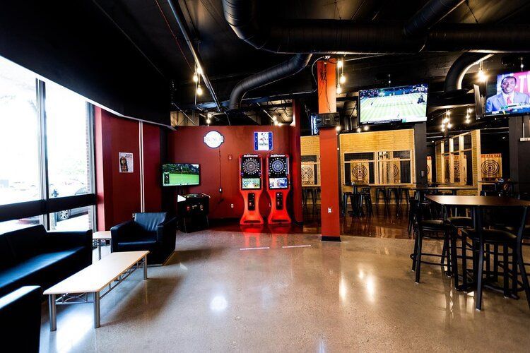 Interior of a modern pub with dartboards, TV screens, black furniture, and red and black walls.