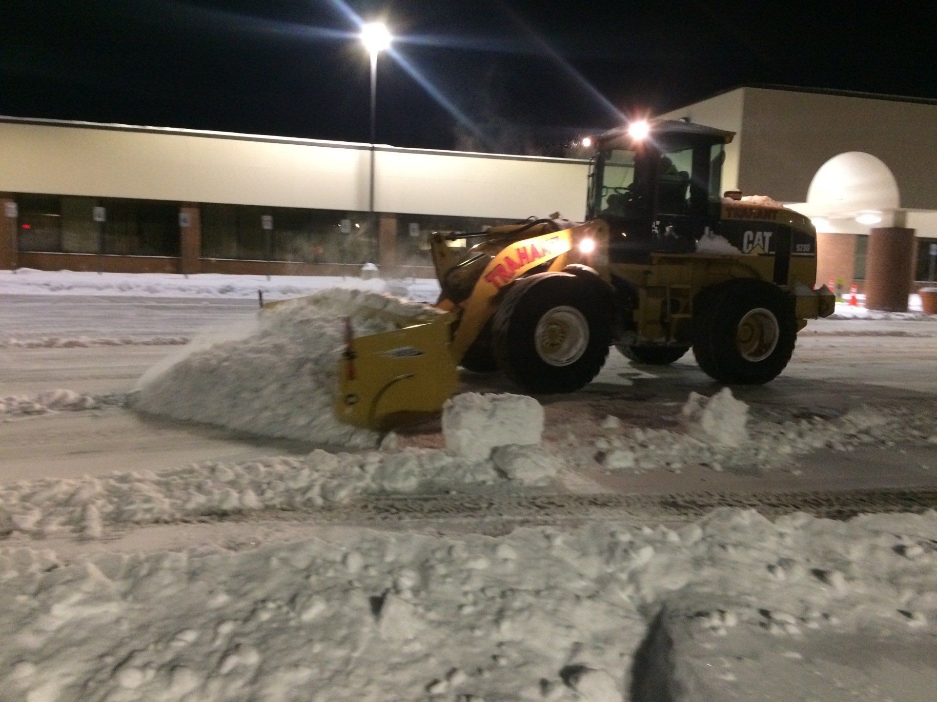 a cat snow plow is clearing snow from a parking lot at night .