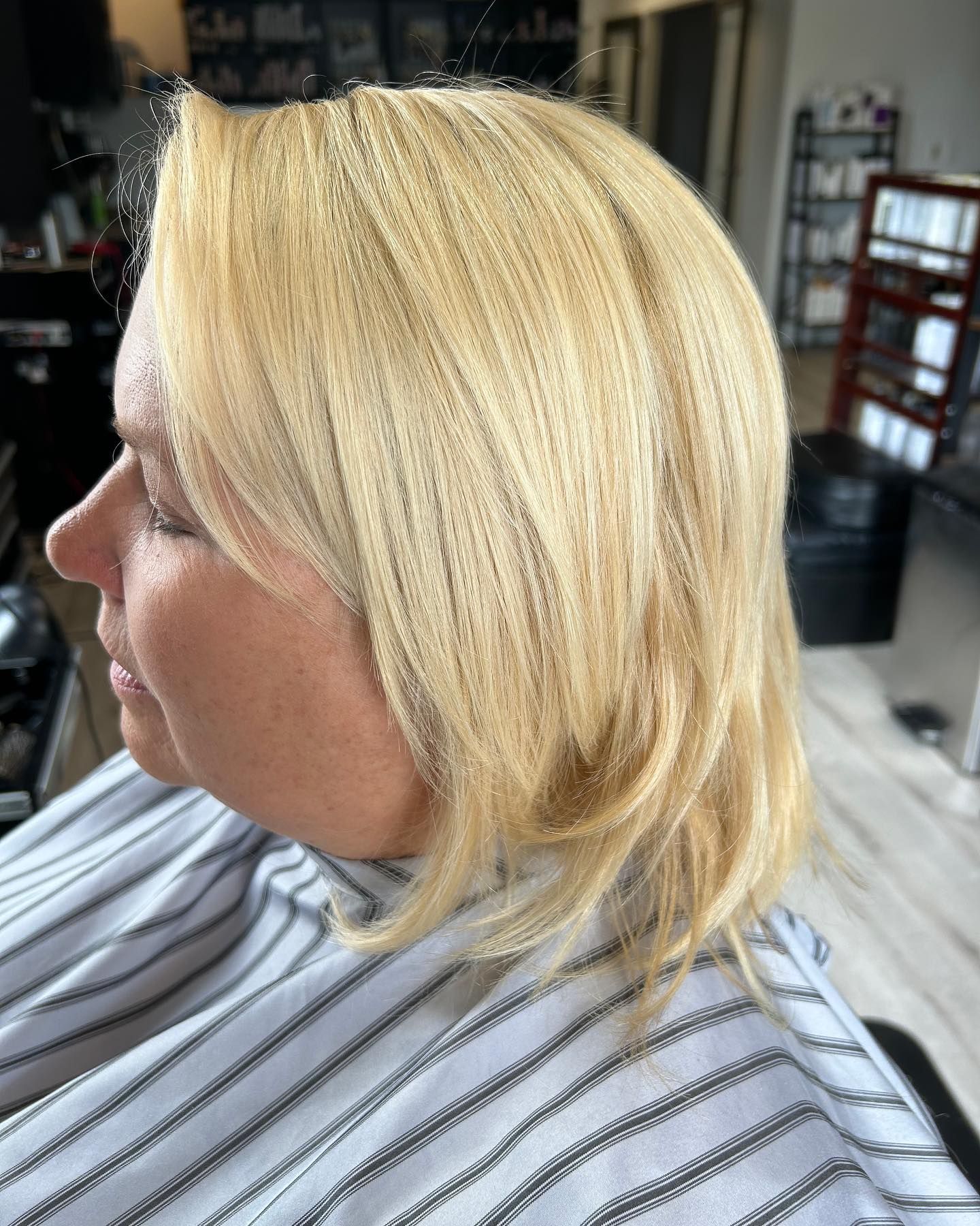 Blonde-haired person, profile view, in a salon chair. Hair is chin-length, styled, light blonde. Wearing a striped salon cape.
