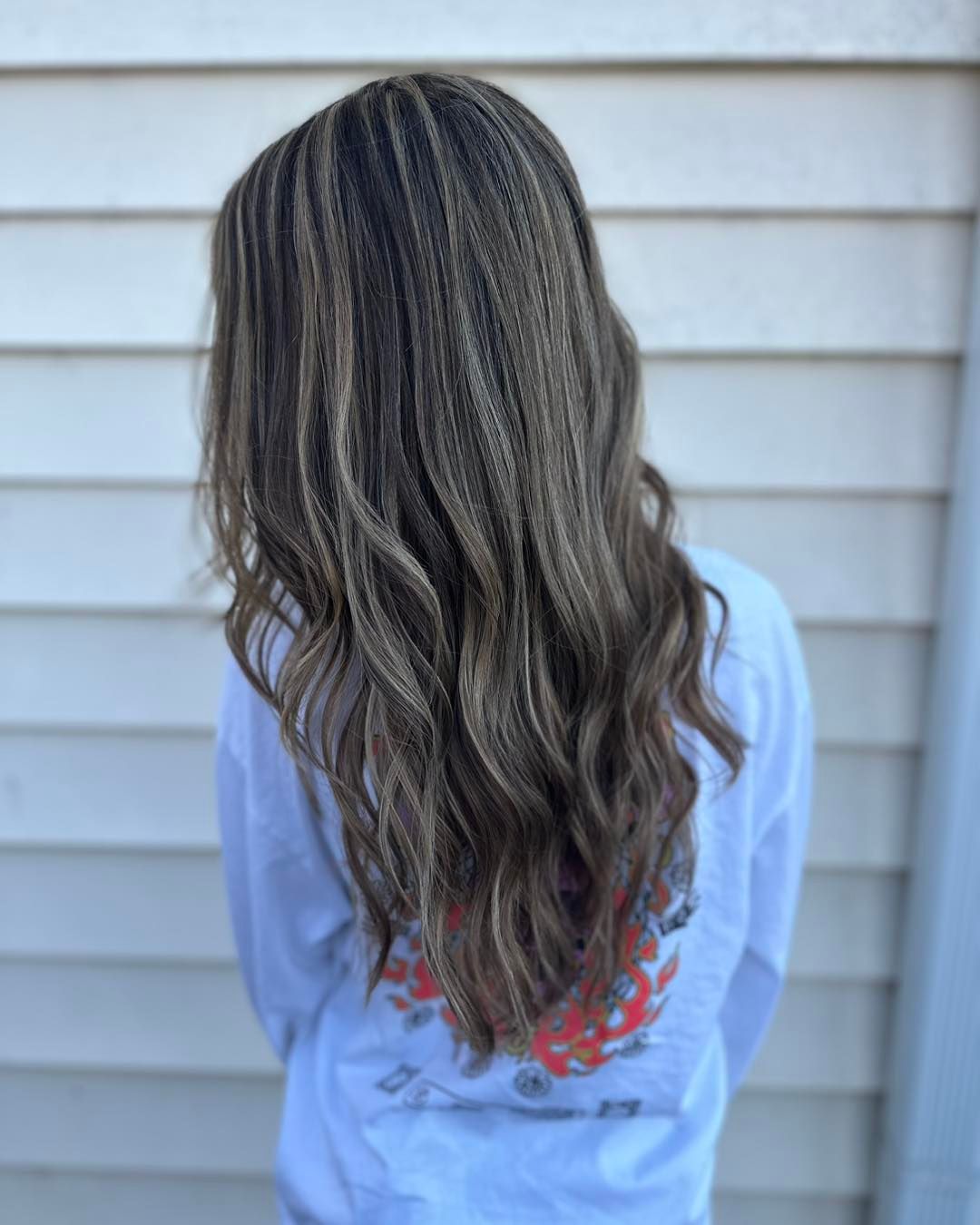 Person with long, crimped, brown and blonde hair, wearing a white shirt, standing outside.