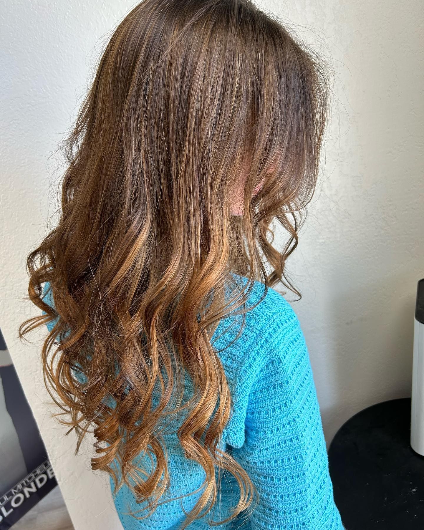 Child with long, wavy brown hair, wearing a blue sweater, in front of a white wall.
