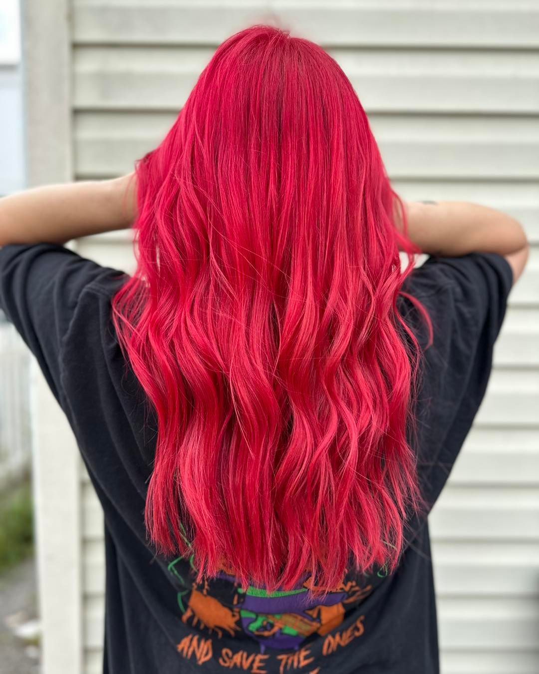 Person with bright red, wavy hair wearing a black t-shirt against a grey wall.