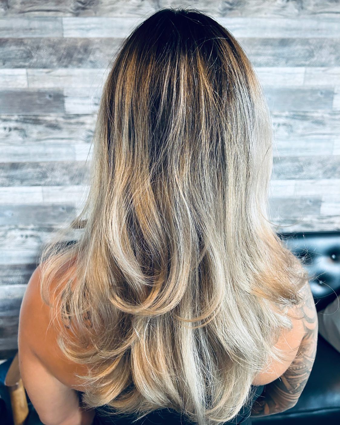 Woman with layered blonde hair, a dark root, and light highlights. She is sitting in front of a gray brick wall.