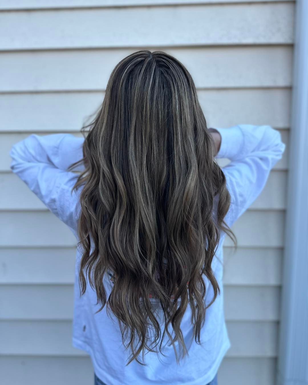 Person with long, crimped, brown and blonde hair, wearing a white shirt, arms raised. Background is white siding.