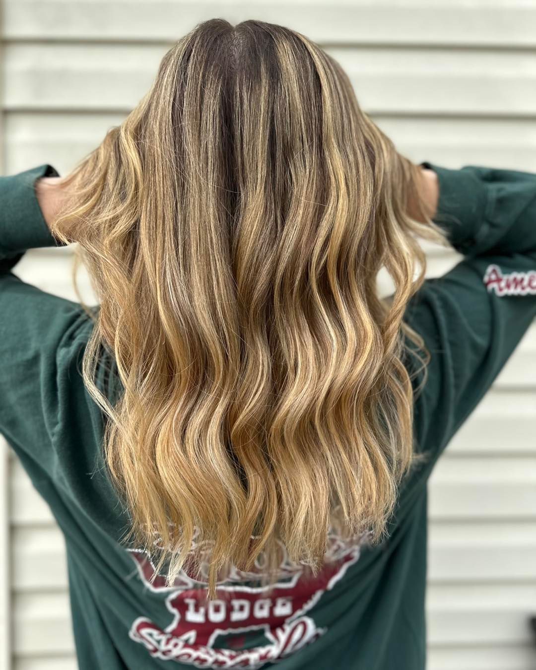Person with wavy, highlighted blonde hair, wearing a green long-sleeved shirt, facing away from the camera.