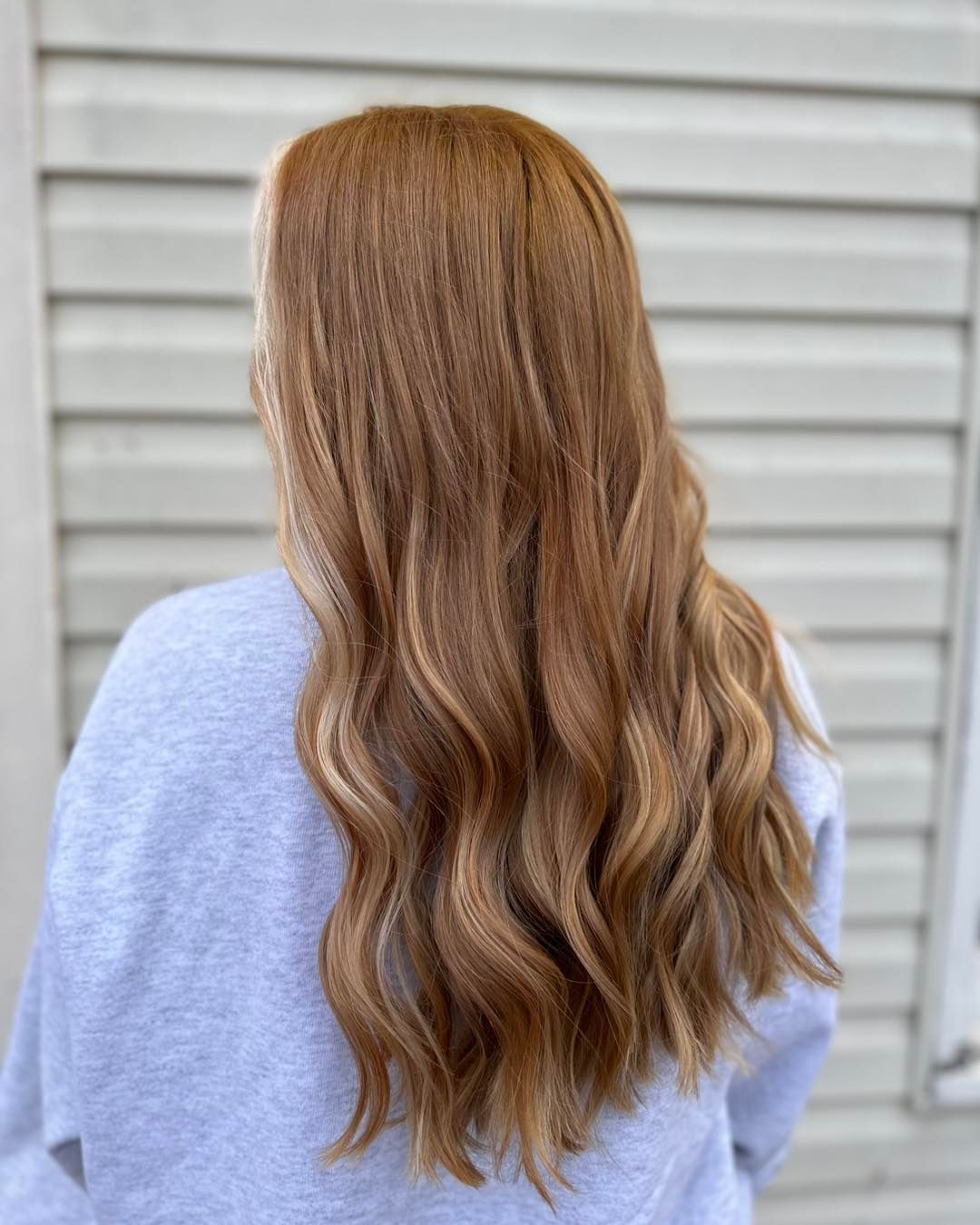Woman with long, wavy auburn hair. Light blonde highlights cascade through the ends. She wears a grey sweatshirt.