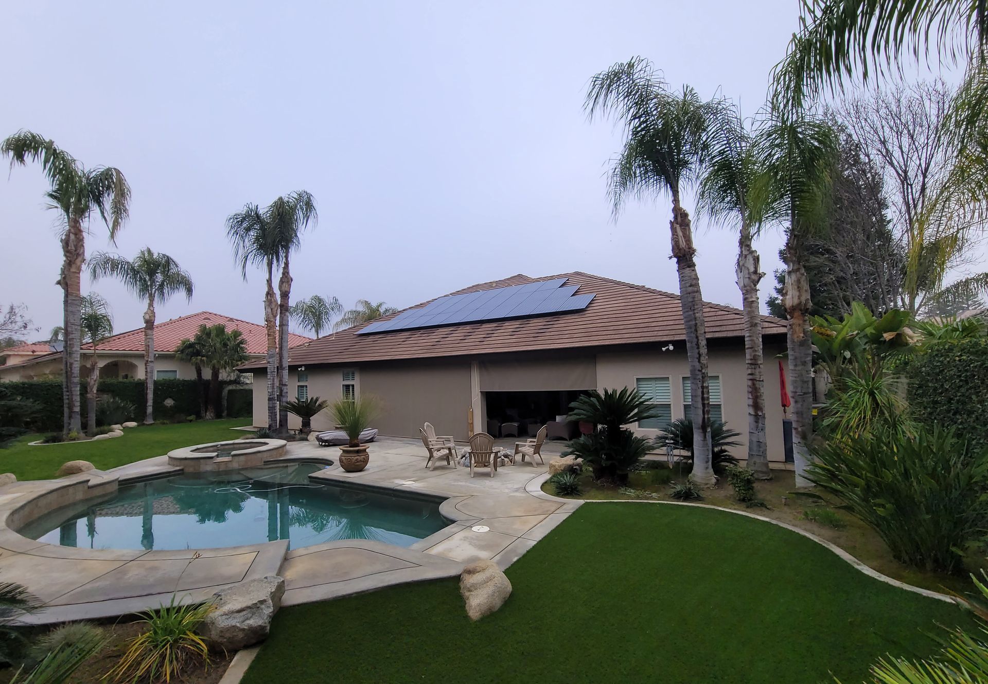 Backyard with pool, lawn, palm trees, and house with solar panels on roof under a cloudy sky.