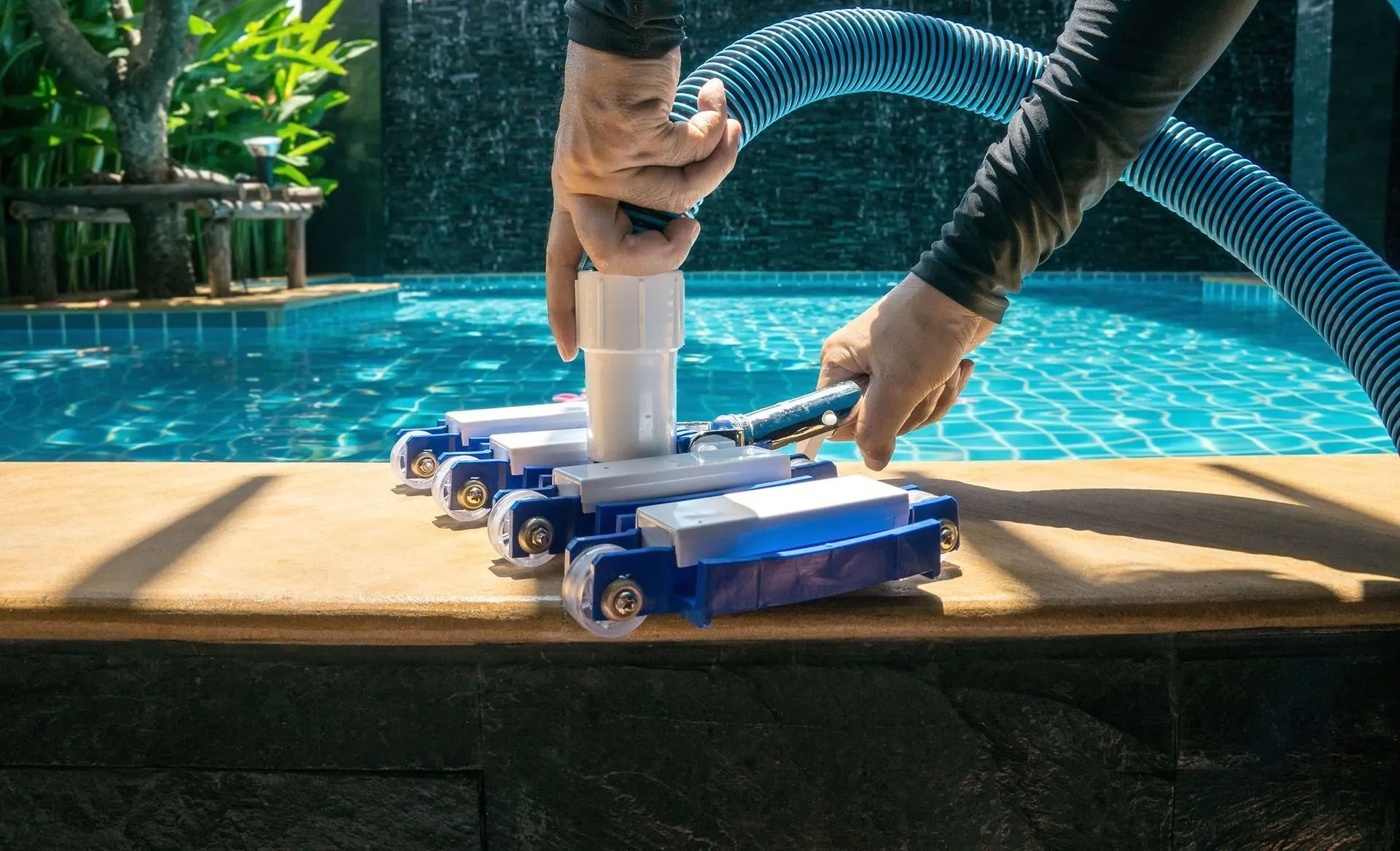 Person attaching pool vacuum to hose near a blue pool.