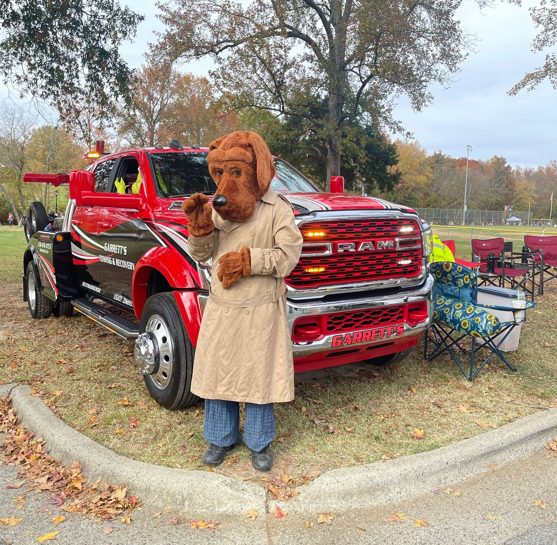 Dog mascot in trench coat stands by a red tow truck, waving. Outdoor setting, fall leaves.