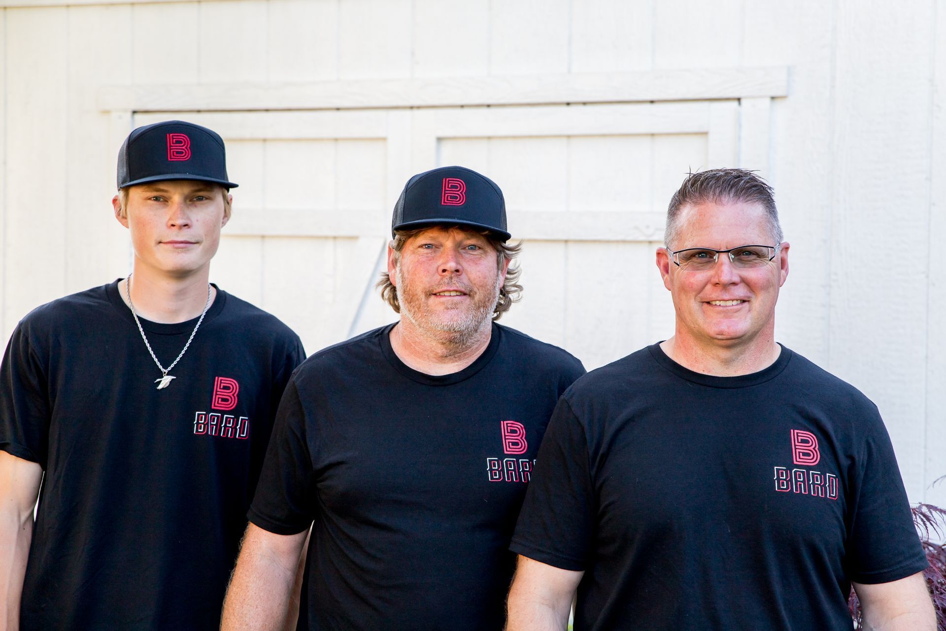 Three men wearing black caps and t-shirts with a red logo stand in front of a white building.
