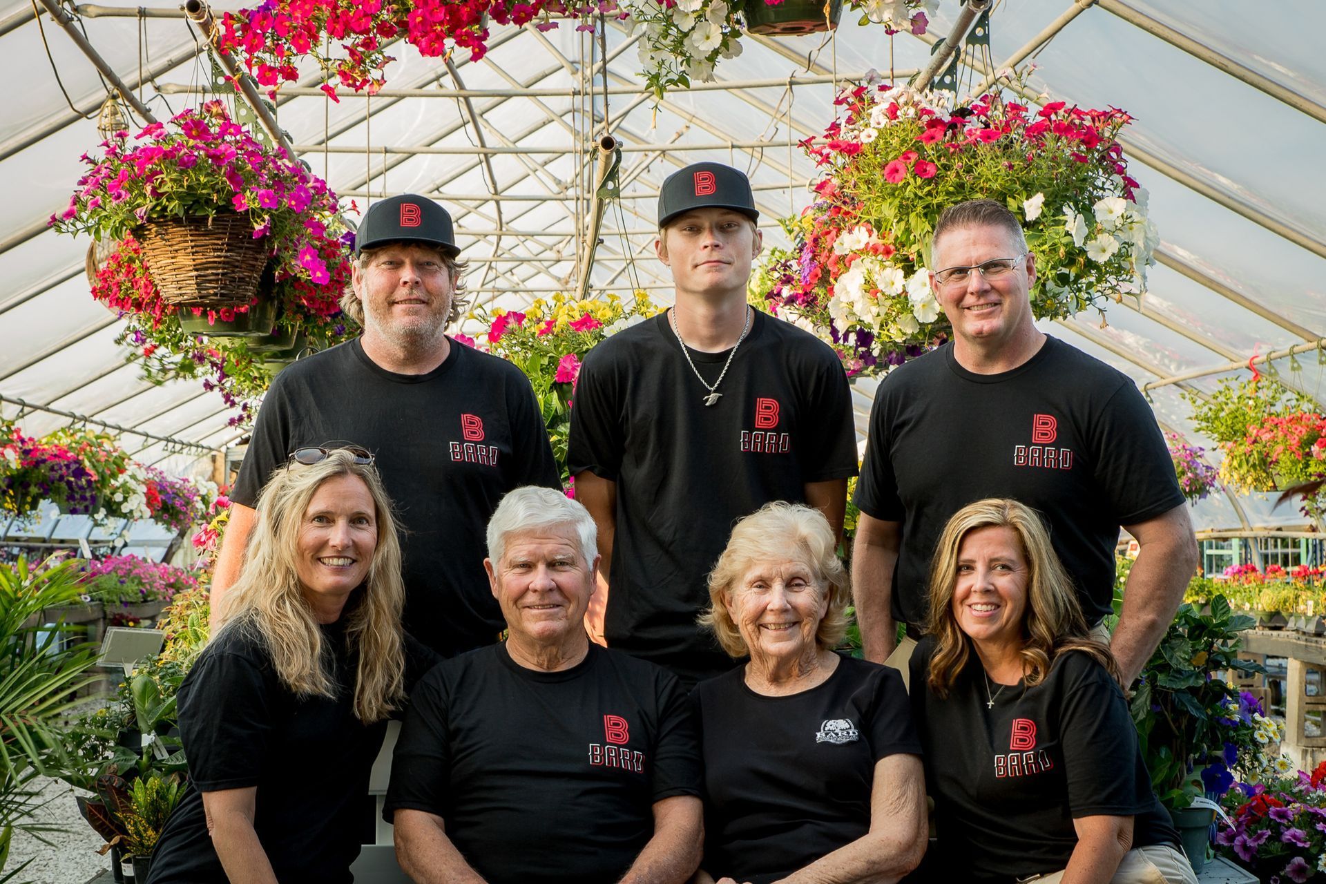 Group of people wearing black shirts in a greenhouse, surrounded by hanging flower baskets.