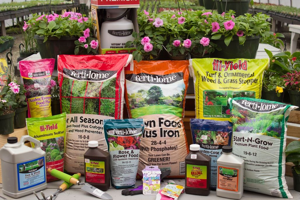 Display of gardening supplies in a greenhouse, including bags of soil, plant food, spray bottles, and potted flowers.