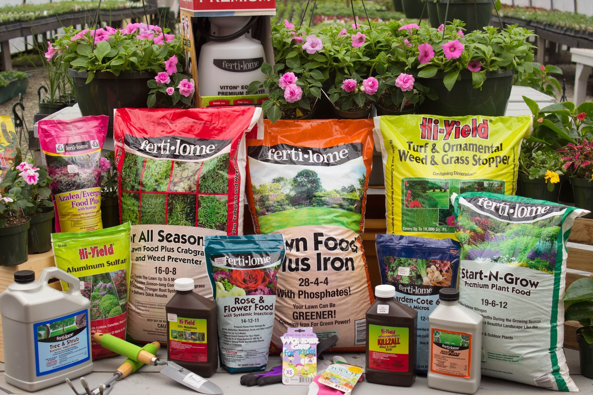 Display of gardening supplies in a greenhouse, including bags of soil, plant food, spray bottles, and potted flowers.