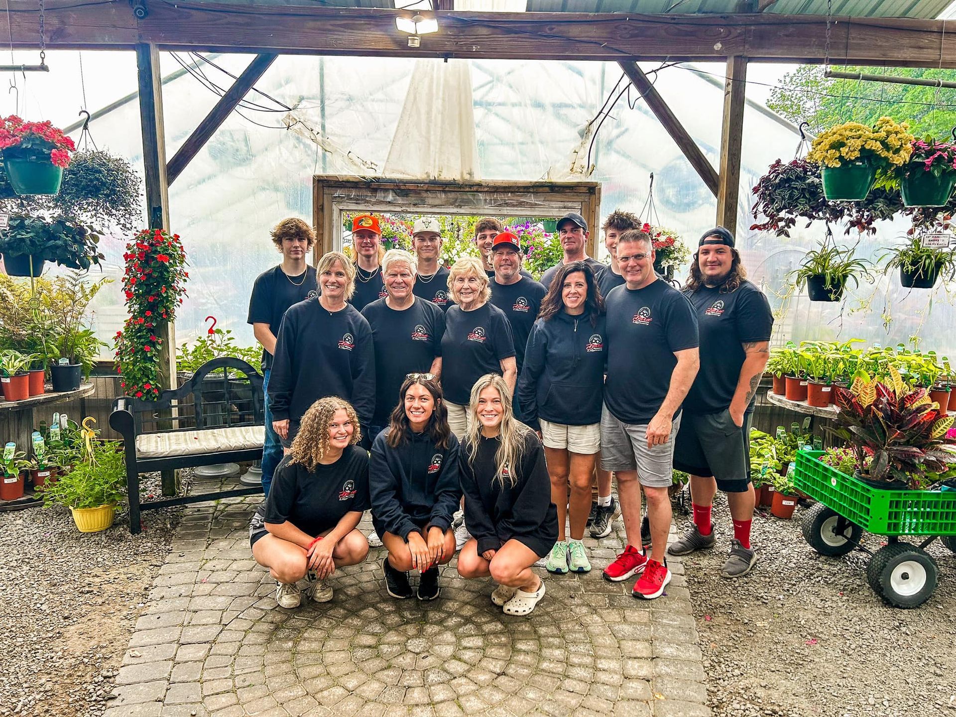 Group of people smiling in a greenhouse, surrounded by plants and flowers; wearing matching shirts.