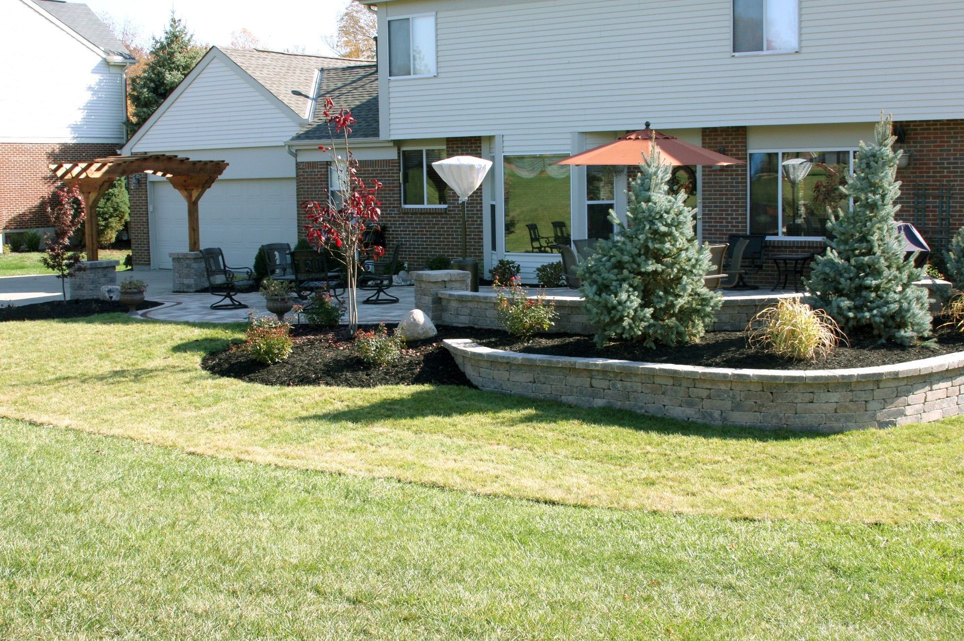 Backyard patio with tiered landscaping and lawn. House in background, trees and a pergola.