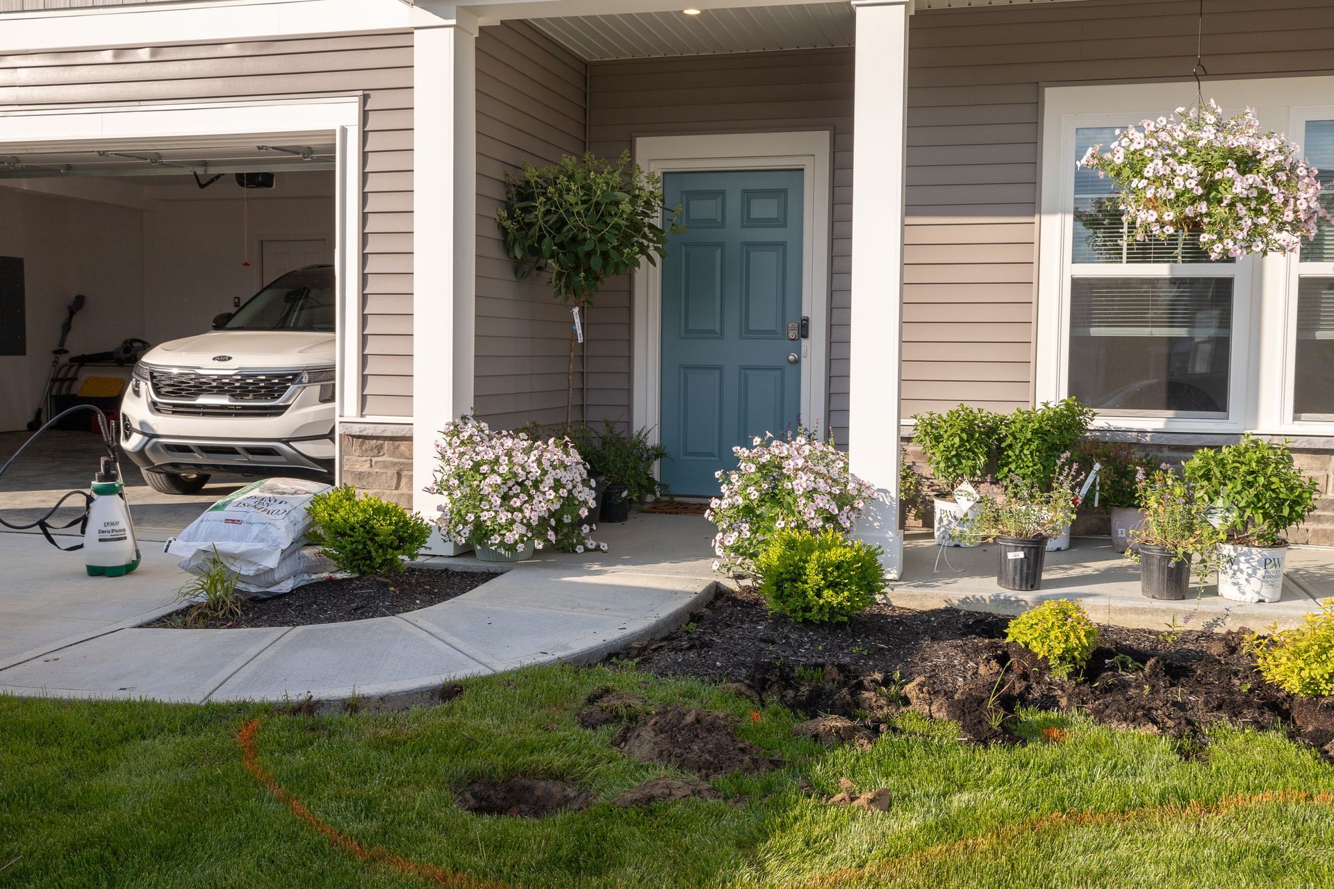 Front porch with landscaping and a car in the garage. Light blue door, hanging flower baskets, and flower beds.