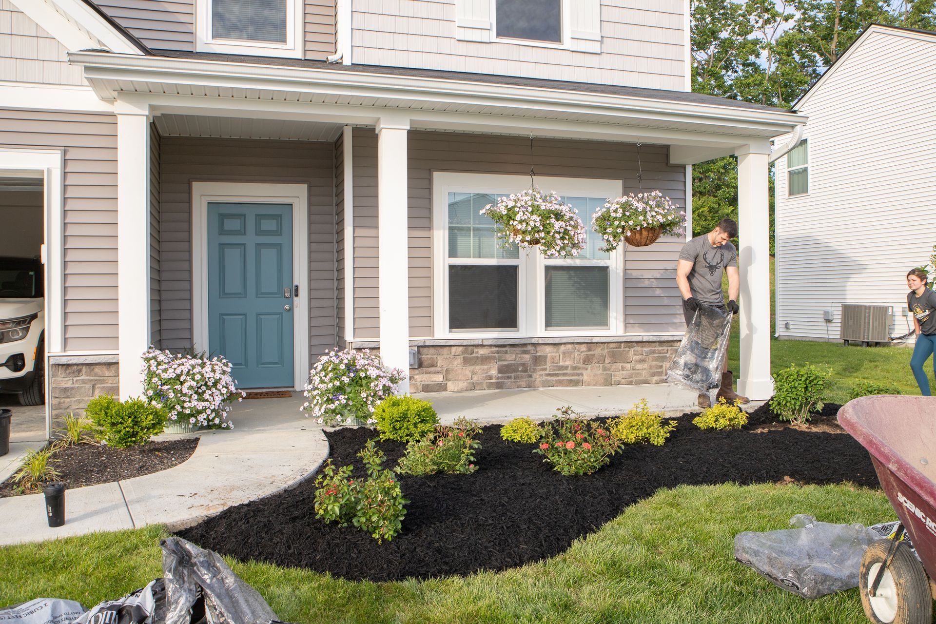 House exterior with porch, landscaping, and people working. Blue door, hanging flower baskets, black mulch, gray siding.