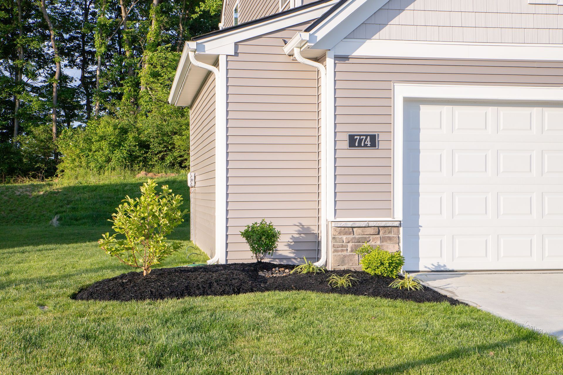 Corner of a house with a garage, tan siding, and a small landscaped bed with mulch and shrubs.