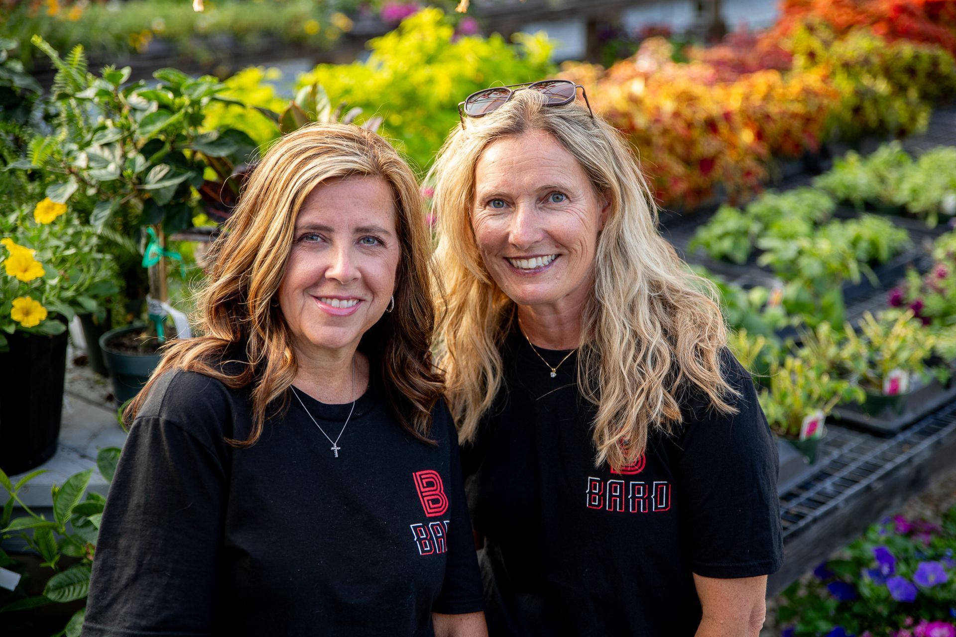 Two women smiling, in a garden, wearing black shirts with logo. Plants in background.