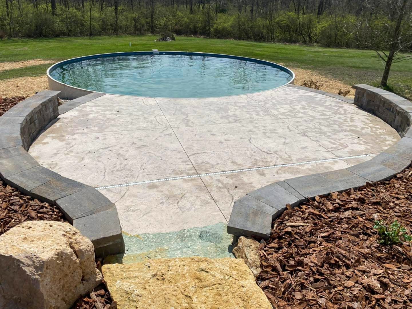 Above-ground pool with concrete patio, rock accents, and curved stone seating in a backyard setting.